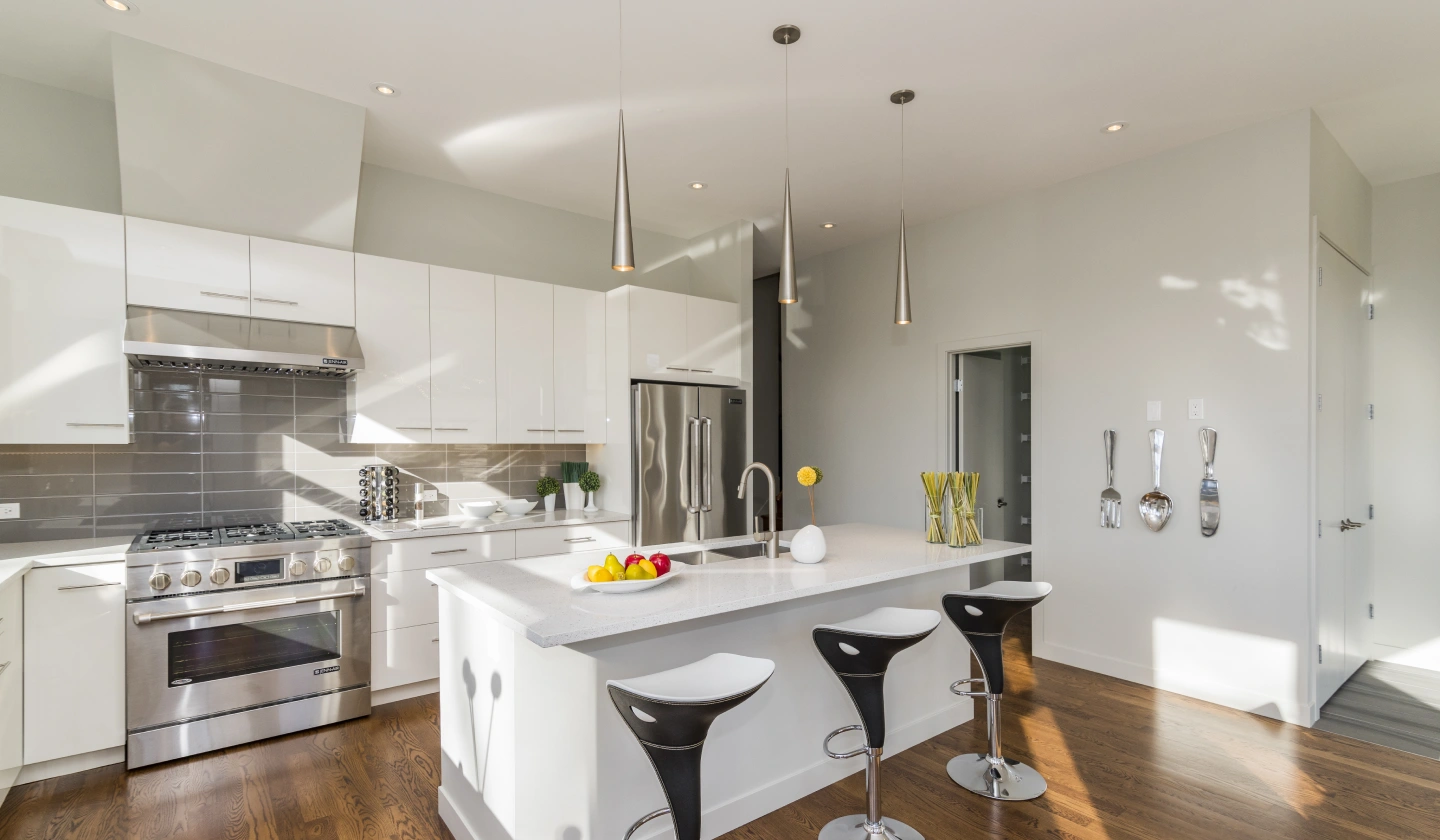 Cocina blanca con isla central iluminada por tres lámparas colgantes y downlights empotrados en el techo
